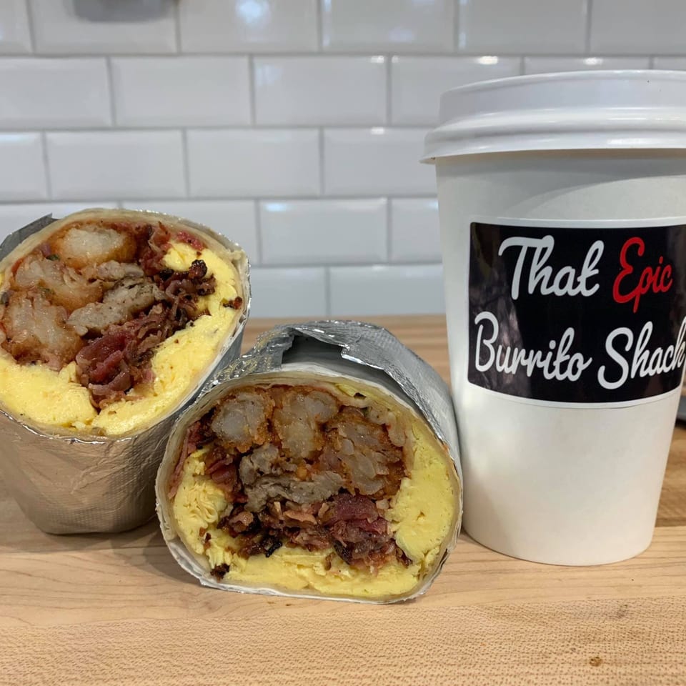 Two foil-wrapped breakfast burritos, cut in half, revealing eggs, bacon, and hash browns, next to a branded coffee cup.