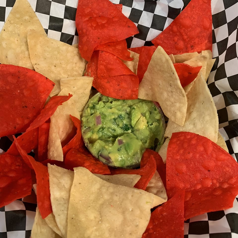 A bowl of guacamole surrounded by red and white tortilla chips in a checkered paper liner.