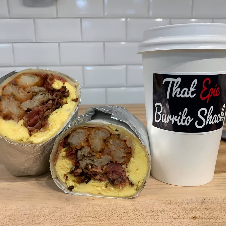 Two breakfast burritos with eggs, bacon, and hash browns, next to a That Epic Burrito Shack cup.