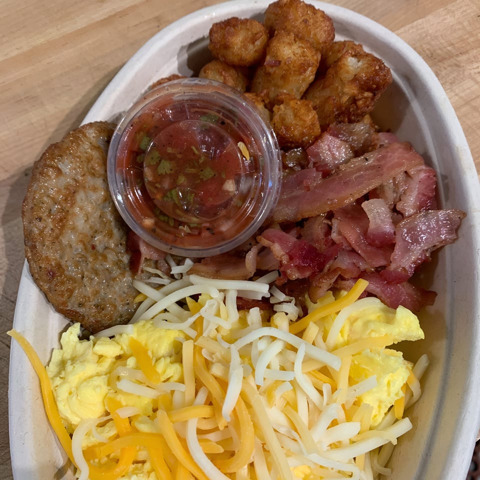 Breakfast bowl with scrambled eggs, cheese, sausage patty, bacon, tater tots, and salsa.